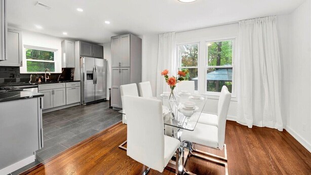 A modern kitchen with white cabinetry, a dining area with a glass table, and a view of the outdoors through large windows. - Modern Comfort in the Heart of NYC (Yonkers)