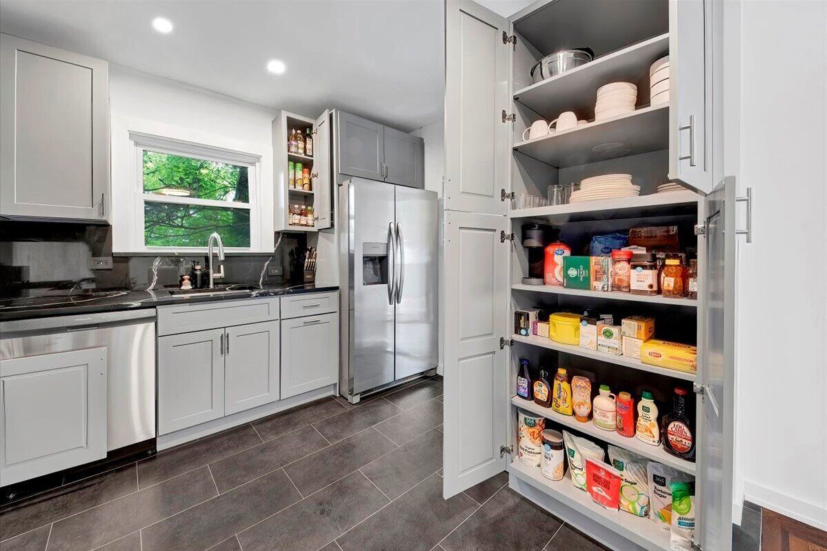 A modern kitchen with grey cabinets, a stainless steel refrigerator, and a pantry stocked with food items.