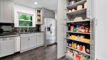A modern kitchen with grey cabinets, a stainless steel refrigerator, and a pantry stocked with food items.