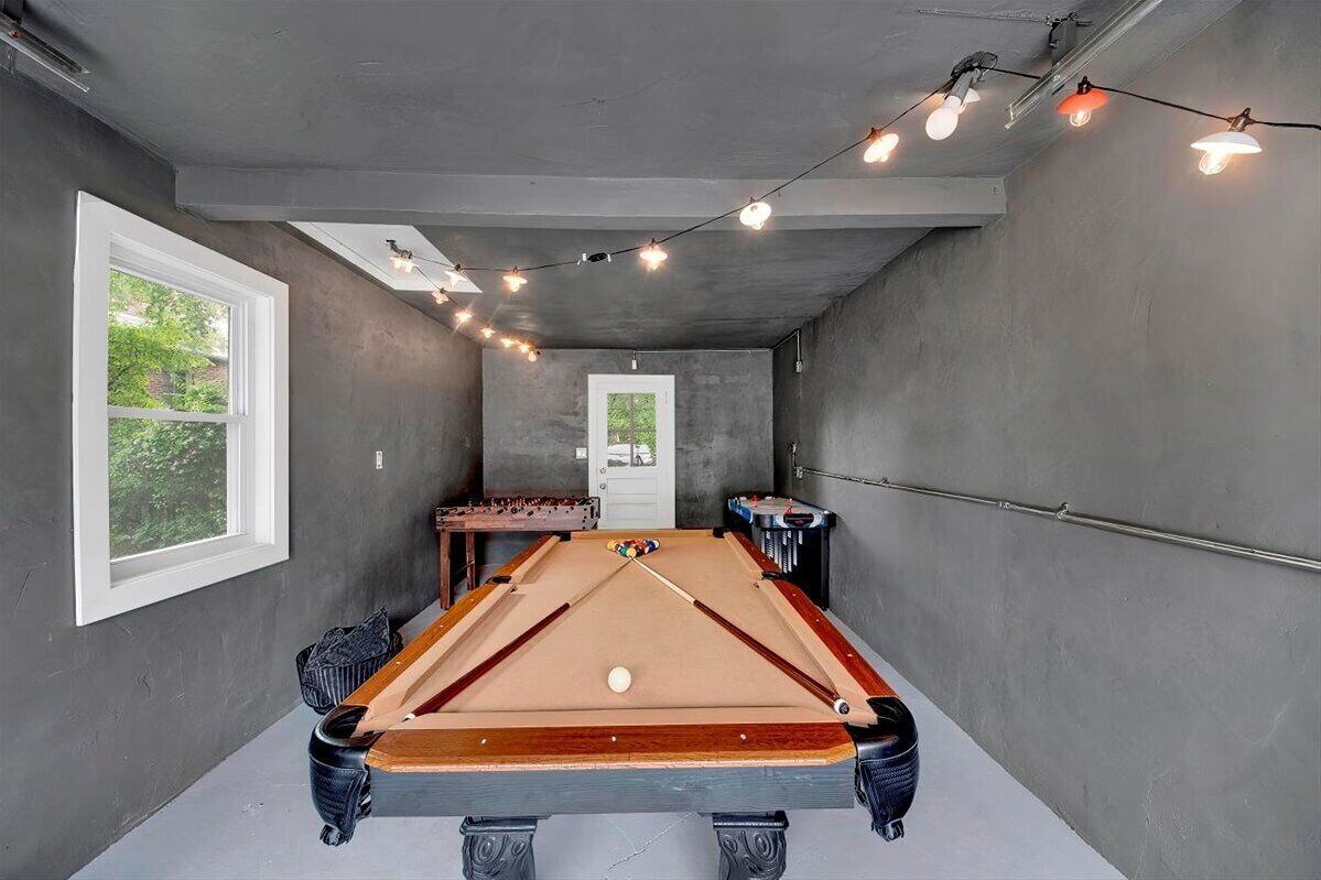 Et billardbord i et rum med betont loft og vægge, et vindue og lyskæder.