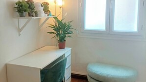 Una habitación pequeña y bien iluminada con un escritorio blanco, una silla verde, un reposapiés redondo pequeño y una ventana con persianas.