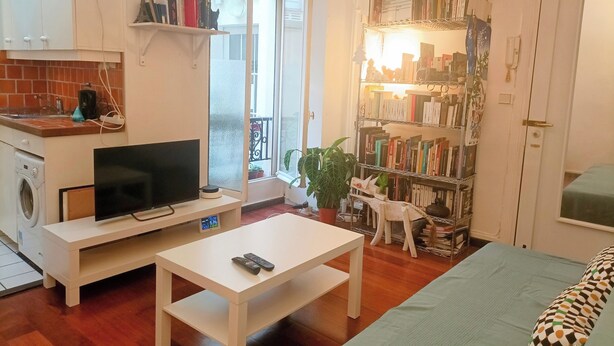 Un espacio habitable compacto con kitchenette, un televisor de pantalla plana, una pequeña mesa, una estantería y un balcón con vistas.