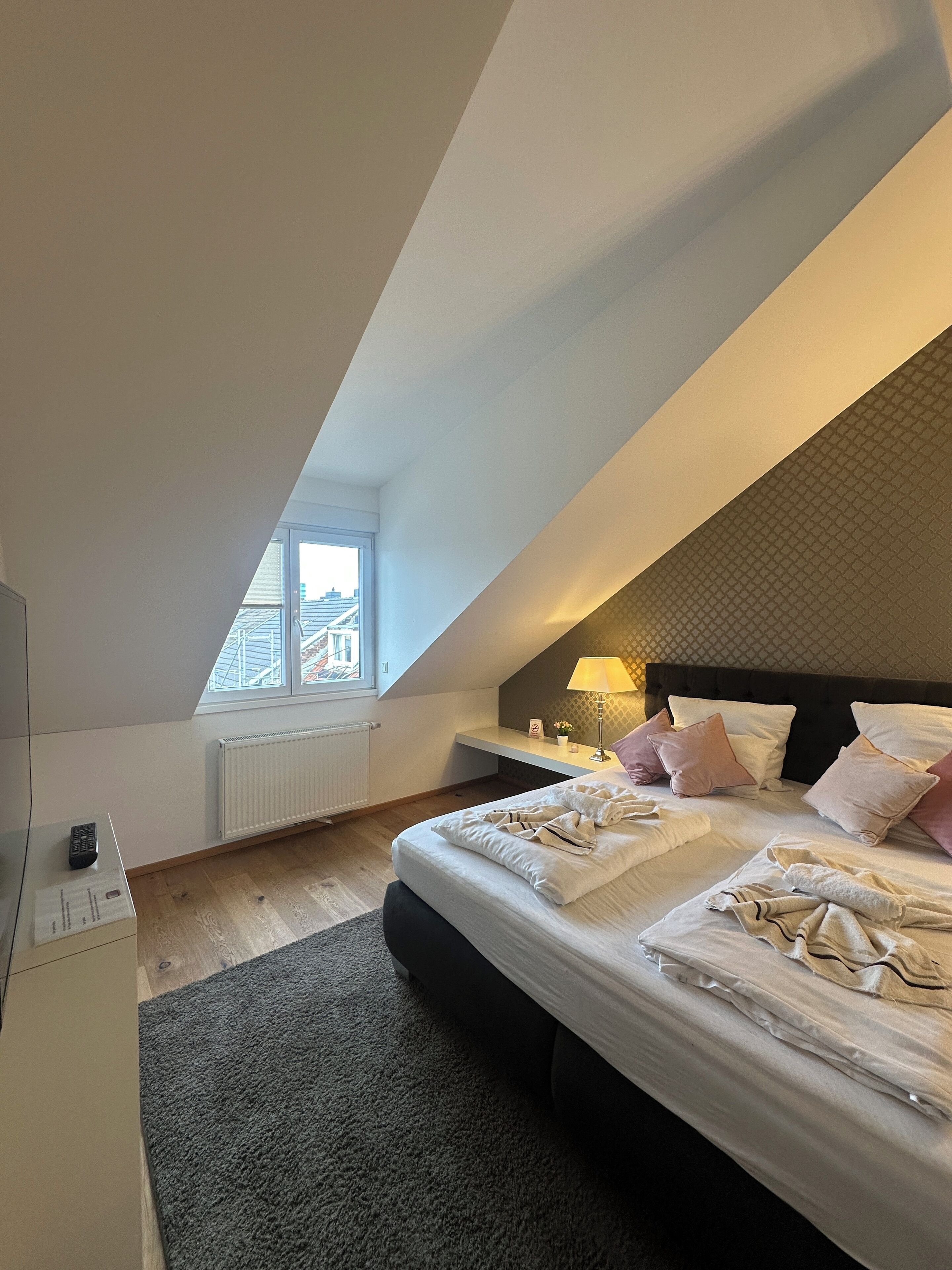 Une chambre avec un plafond en pente, un lit avec des oreillers et une couverture, une table de chevet avec une lampe, une fenêtre donnant sur des immeubles et un radiateur.