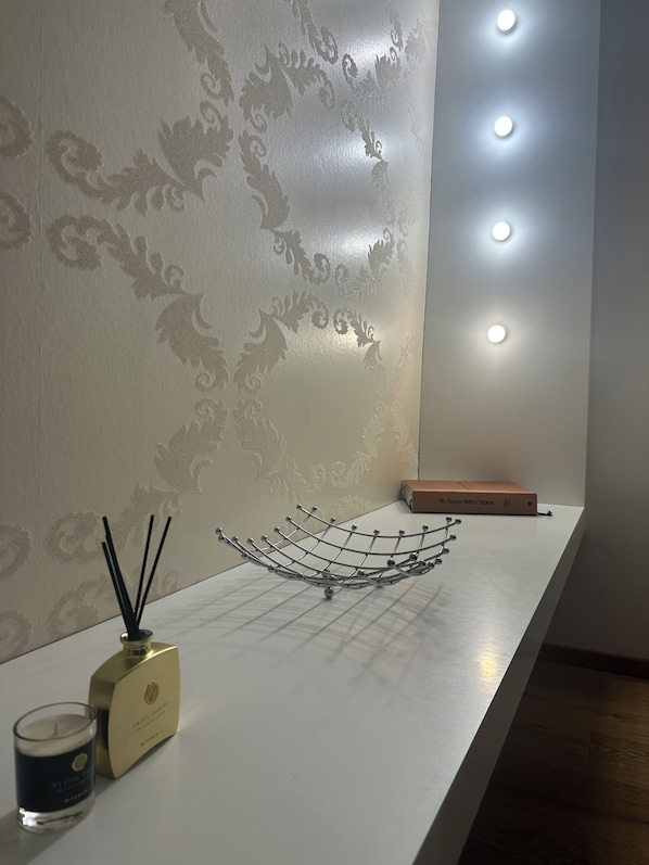 A room with a decorative wall, a white shelf with a candle, a perfume bottle, and a book. - Haven - Helvetia Suites Düsseldorf City (Düsseldorf)