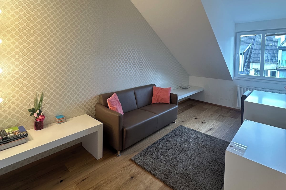 A modern loft bedroom with a sloped ceiling, a brown leather sofa, a white bench, and a large window offering a city view.