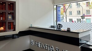 A hotel reception area with a modern design, featuring a reception desk, a digital display, and a view of the street outside. - Haven - Helvetia Suites Düsseldorf City (Düsseldorf)