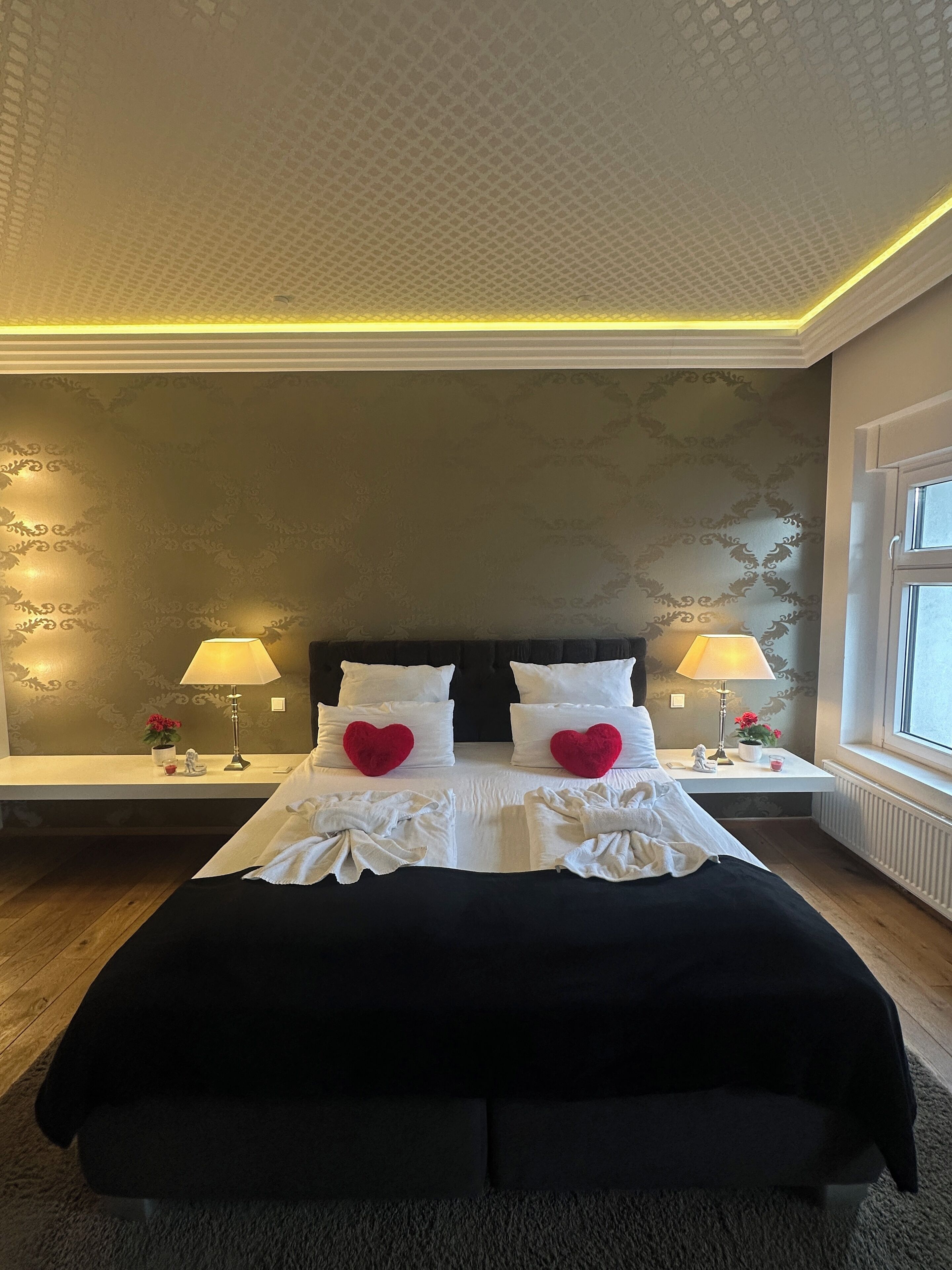 A hotel room with a large bed, two bedside tables with lamps, a window with a view, and a patterned wallpaper.