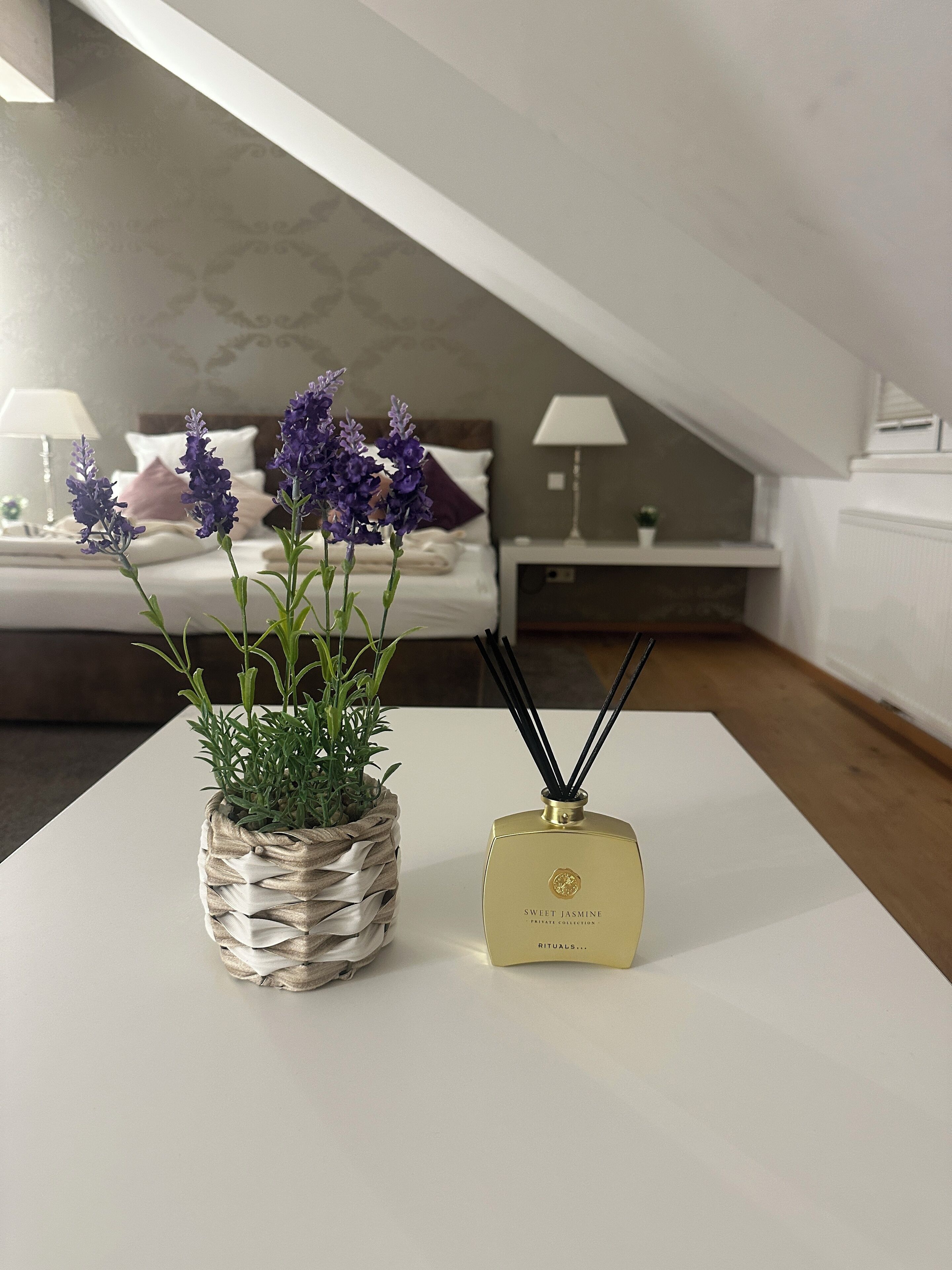 A bedroom with a bed, a bedside table with a lamp, a small potted plant, and a diffuser on a white table.