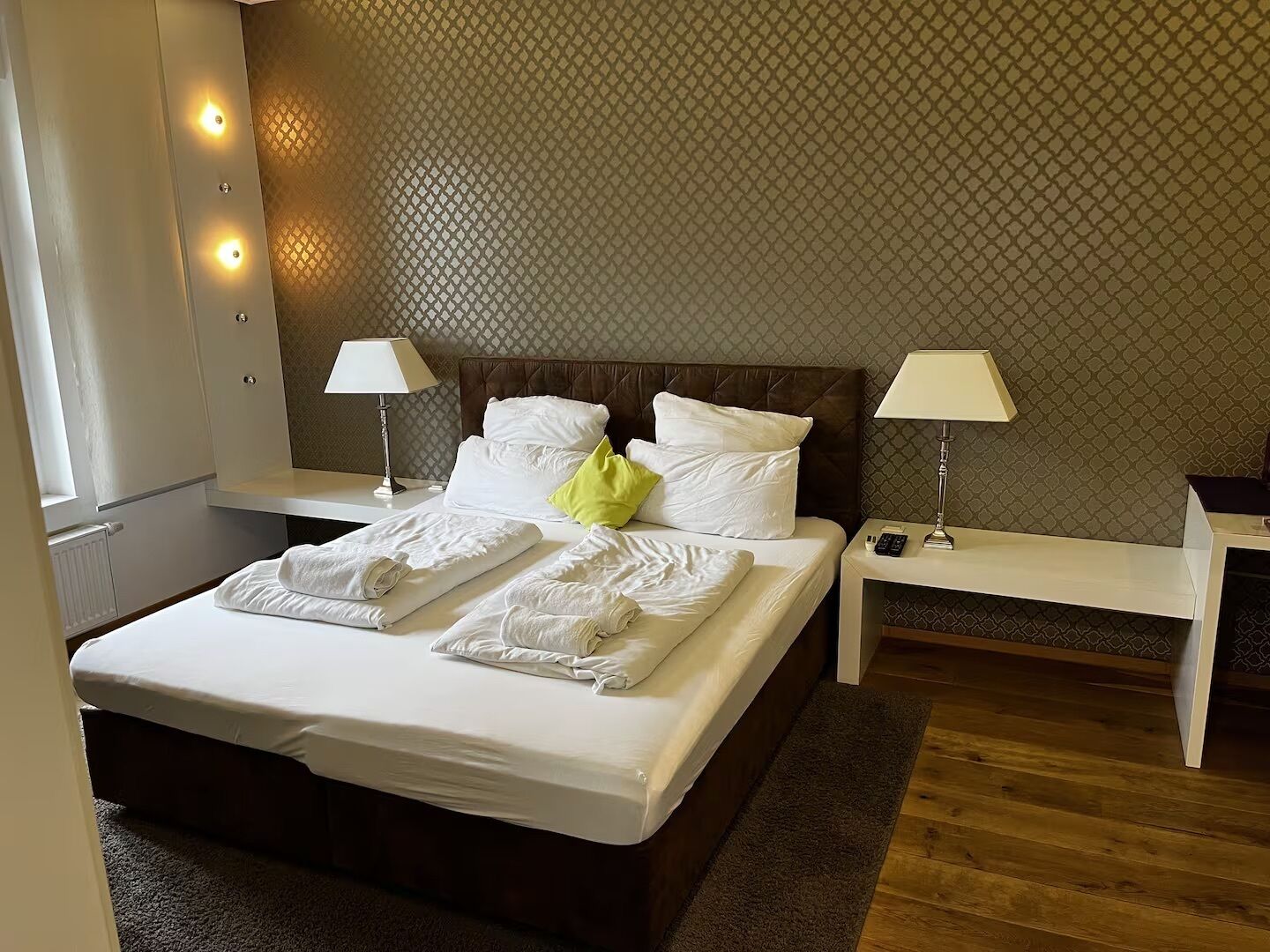 A neatly made bed with white linens and a green pillow, two bedside lamps, and a patterned wall.