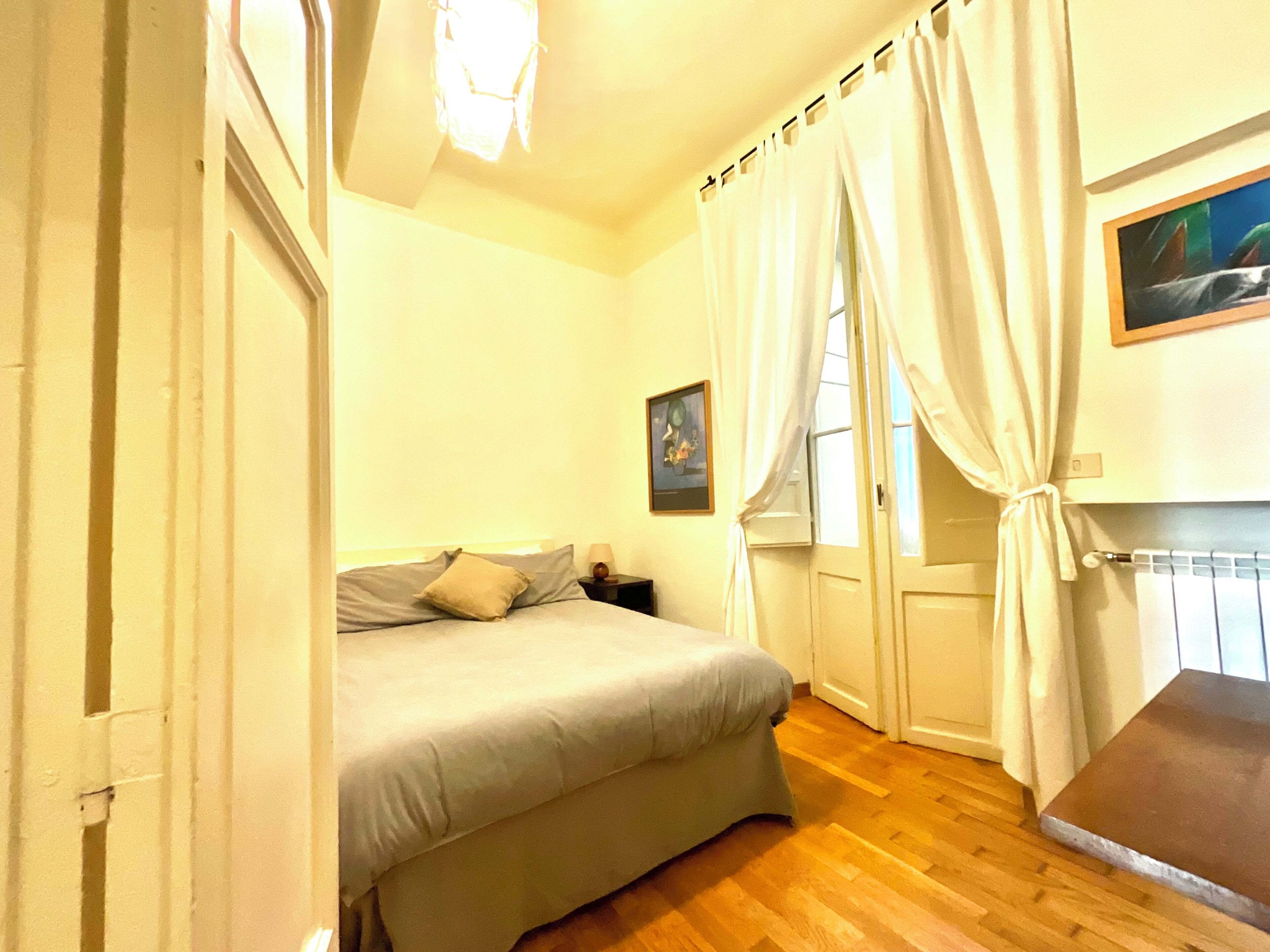 Ein Schlafzimmer mit Bett, Nachttisch, Fenster mit Vorhängen und einem Bild an der Wand.