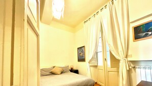 A bedroom with a bed, a nightstand, a window with curtains, and a painting on the wall. - Spanish Steps from my window (ROMA)