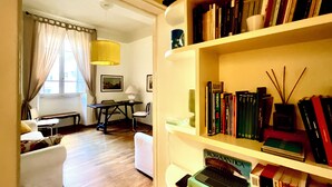 A room with a wooden floor, a white sofa, a dining table with chairs, a bookshelf with books, and a window with curtains. - Spanish Steps from my window (ROMA)