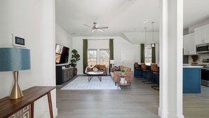 A spacious living area with a sofa, coffee table, and a TV. A kitchen with a blue countertop and cabinets is visible in the background. - Relax, Work, Play! Charming 3-bedroom house in amazing Dallas with Game Room (Dallas)