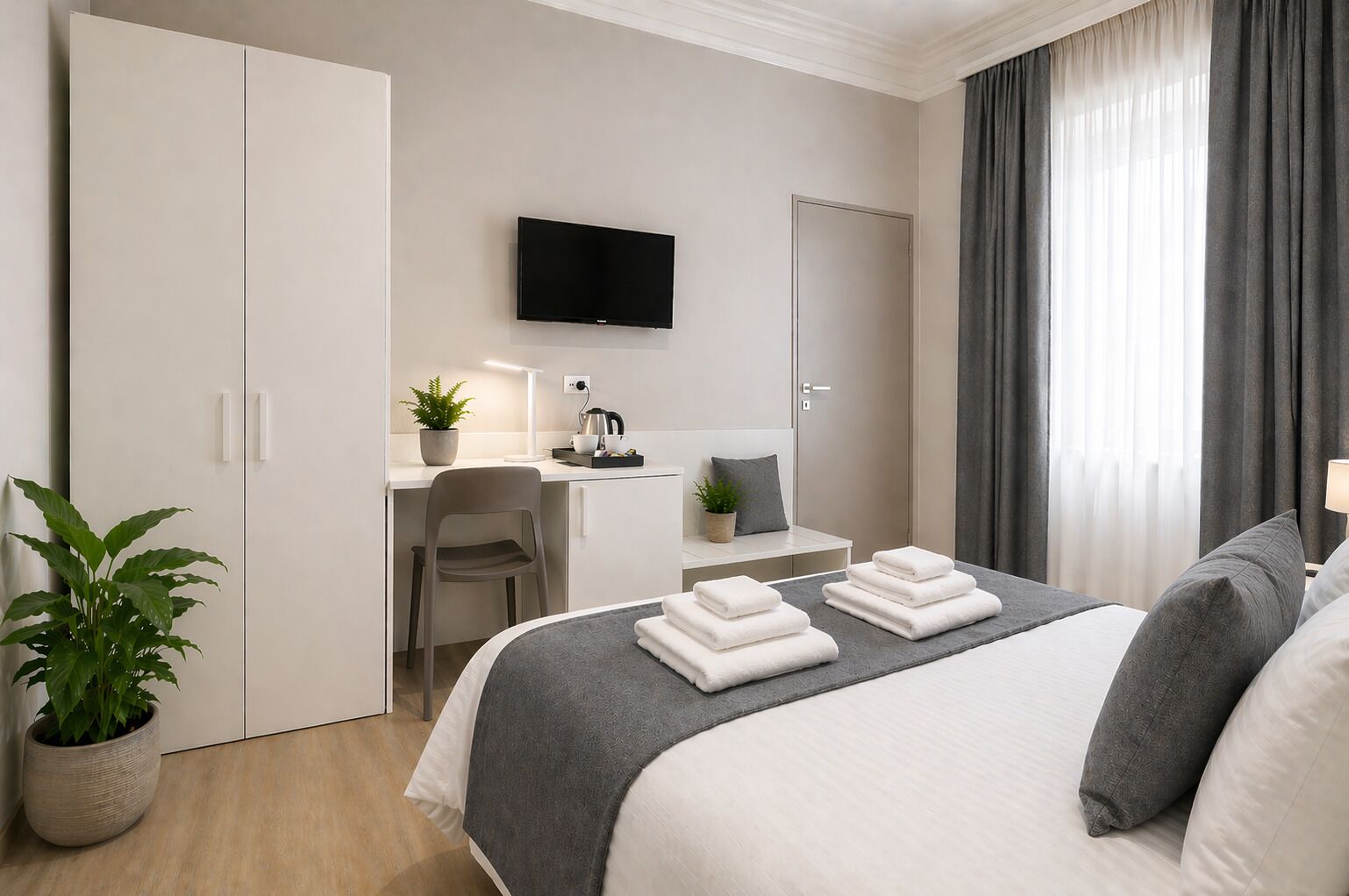 Habitación de hotel con cama, escritorio, televisor de pantalla plana, planta en maceta y ventana grande con cortinas.