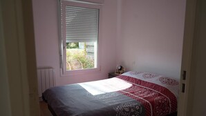 A bedroom with a bed, a window with blinds, a radiator, and a small bedside table with a lamp. - Quiet, warm apartment (Miniac-Morvan)