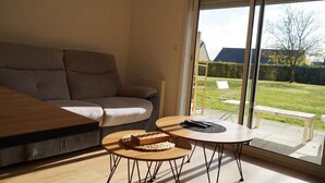 A modern living room with a sofa, a coffee table, and a view of a garden through a sliding glass door. - Quiet, warm apartment (Miniac-Morvan)