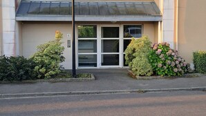 A building with a beige facade, a flat roof, and a small entrance with glass doors. There are two small windows on the upper floor and a single window on the ground floor. A street lamp is visible in front of the entrance. - Quiet, warm apartment (Miniac-Morvan)
