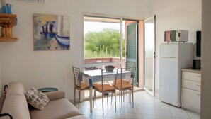 A living room with a sofa, a dining table with chairs, a kitchen area with a refrigerator, and a sliding glass door leading to an outdoor area. - BouganVilla - Two-room apartment with sea view in Badesi (Badesi)