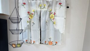 A room with a white curtain featuring floral embroidery, a black hanging basket with a fruit, and a wall socket.
