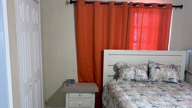 A bedroom with a bed, a nightstand, a door, and an orange curtain.