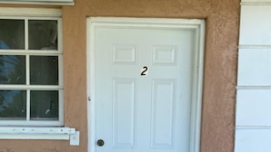 A white door with the number 2, a window with blinds, and a concrete ground.