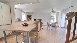 A spacious kitchen with a wooden table, modern chairs, and a fruit bowl. There is a dining area with a table and chairs, and a kitchen island with a sink and a wooden cabinet.