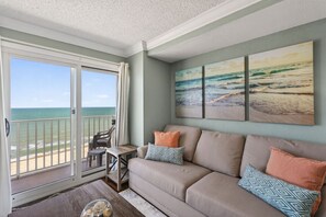 Un salon avec un balcon donnant sur l’océan, un canapé beige et trois œuvres d’art murales sur le thème de la plage.