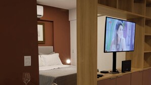 Une chambre d’hôtel avec un lit, un téléviseur posé sur un meuble en bois, une table à manger avec une chaise, et une petite table avec un verre à vin et une assiette.