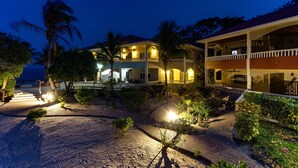 A two-story hotel with a covered outdoor area, palm trees, and a well-lit pathway. - 2BR Beachfront Villa w/ Pool, Sea View and Beach (Placencia)