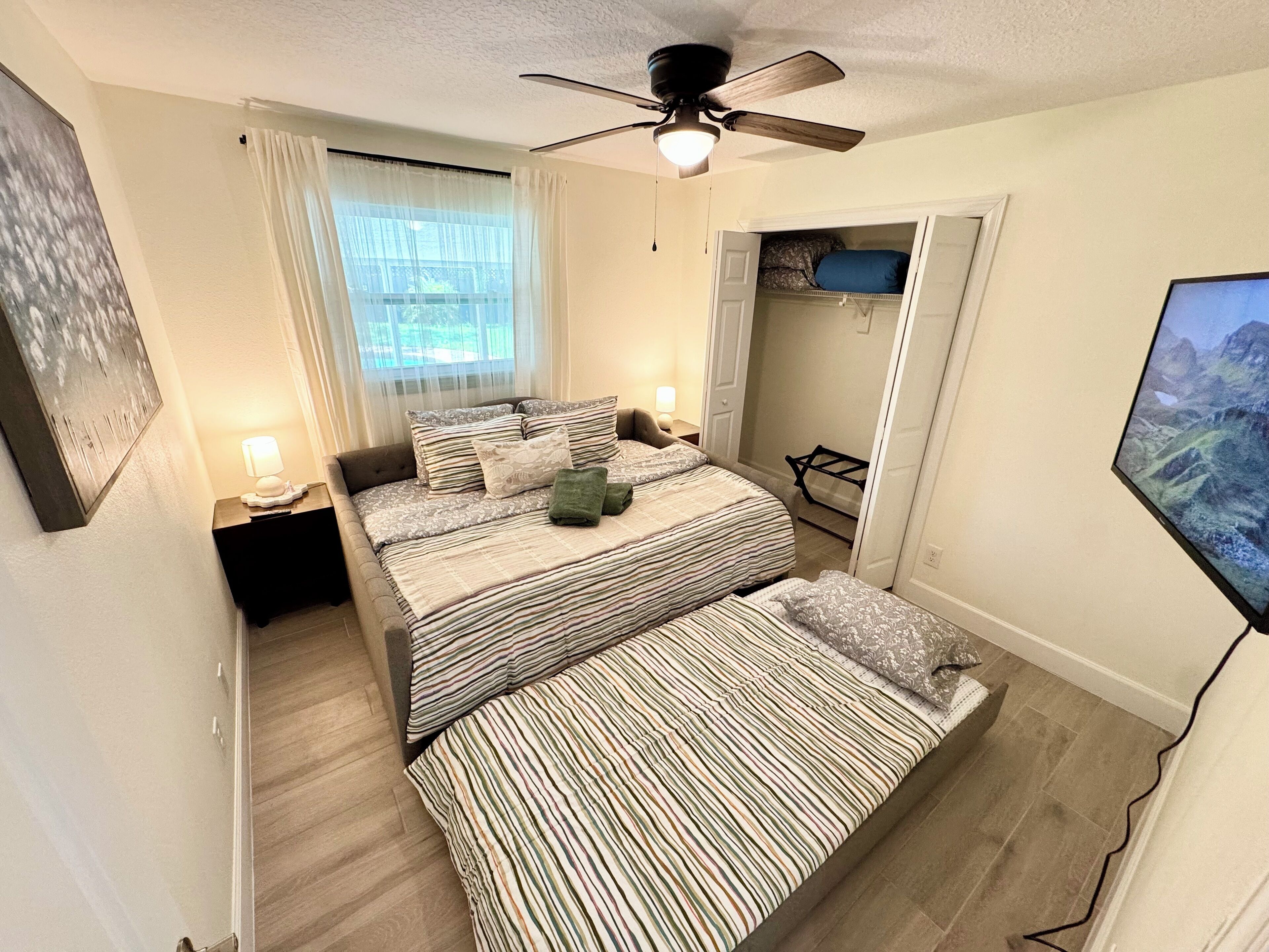 A bedroom with a bed, a ceiling fan, a window with curtains, and a TV mounted on the wall.