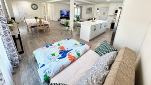 A modern living room with a sofa, a dining area with a table and chairs, a kitchen with a white countertop, and a TV in the background.