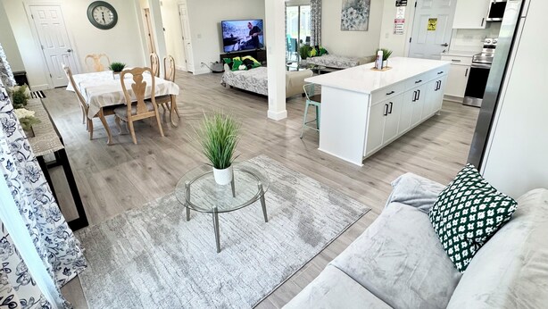 A modern living room with a sofa, a dining area with a table and chairs, a kitchen with white cabinets, and a TV in the background.
