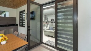 A modern kitchen with a wooden ceiling, a dining area with a table and chairs, and a view into a living room through a glass door.
