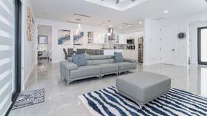 A modern living room with a grey sofa, a grey ottoman, and a blue and white striped rug. There is a ceiling fan, recessed lighting, and a kitchen in the background.
