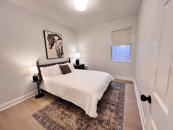 A bedroom with a bed, two bedside tables, a nightstand, a rug, a window, and a painting on the wall.