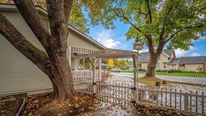 A covered porch with a white fence, a tree with autumn leaves, and a residential street. - Charming Downtown Home, Walk to CDA Shops&Eateries (Coeur d'Alene)