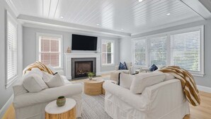 Ein geräumiges Wohnzimmer mit weißen Sofas, einem Kamin und großen Fenstern mit Jalousien.