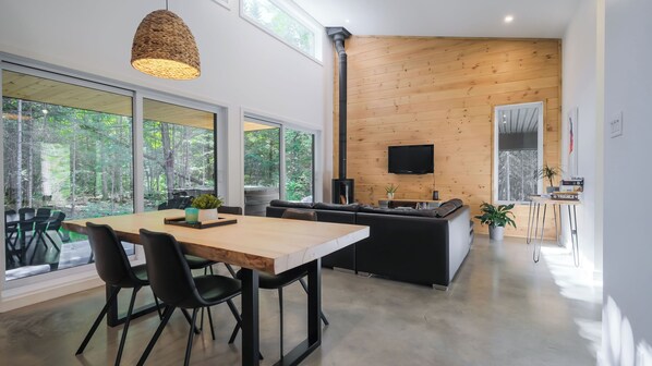 Une cuisine moderne dotée d’une cuisinière intégrée, un coin repas avec une table et des chaises en bois, et de grandes fenêtres offrant une vue sur la forêt.
