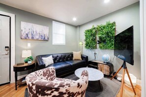 A modern living room with a black leather sofa, a patterned armchair, a round coffee table, a flat-screen TV on a tripod stand, a green wall-mounted plant, and a cityscape painting. - The Bucktown Nook | By Transit w/ Free Parking (Chicago)