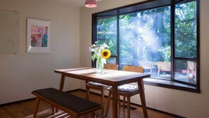Un coin repas avec une table et un banc en bois, un vase rempli de fleurs et une grande fenêtre donnant sur les arbres.