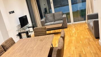 A modern living room with a wooden dining table, sofas, and a large sliding glass door leading to a balcony.