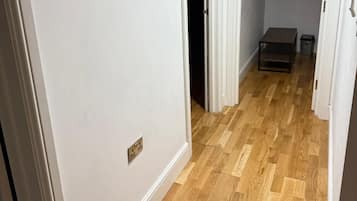 Un couloir doté d’un plancher en bois, de murs blancs et d’une porte donnant sur une pièce sombre.