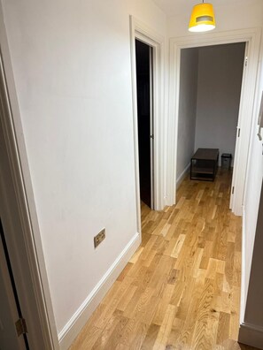 Un pasillo con piso de madera, paredes blancas y una puerta que da a una habitación oscura.
