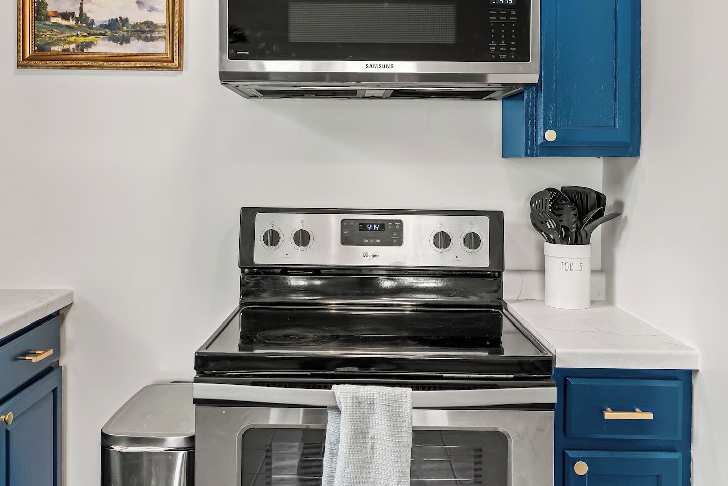 A kitchen with a stainless steel microwave, a Whirlpool stove, and an oven. There are blue cabinets and a white countertop.
