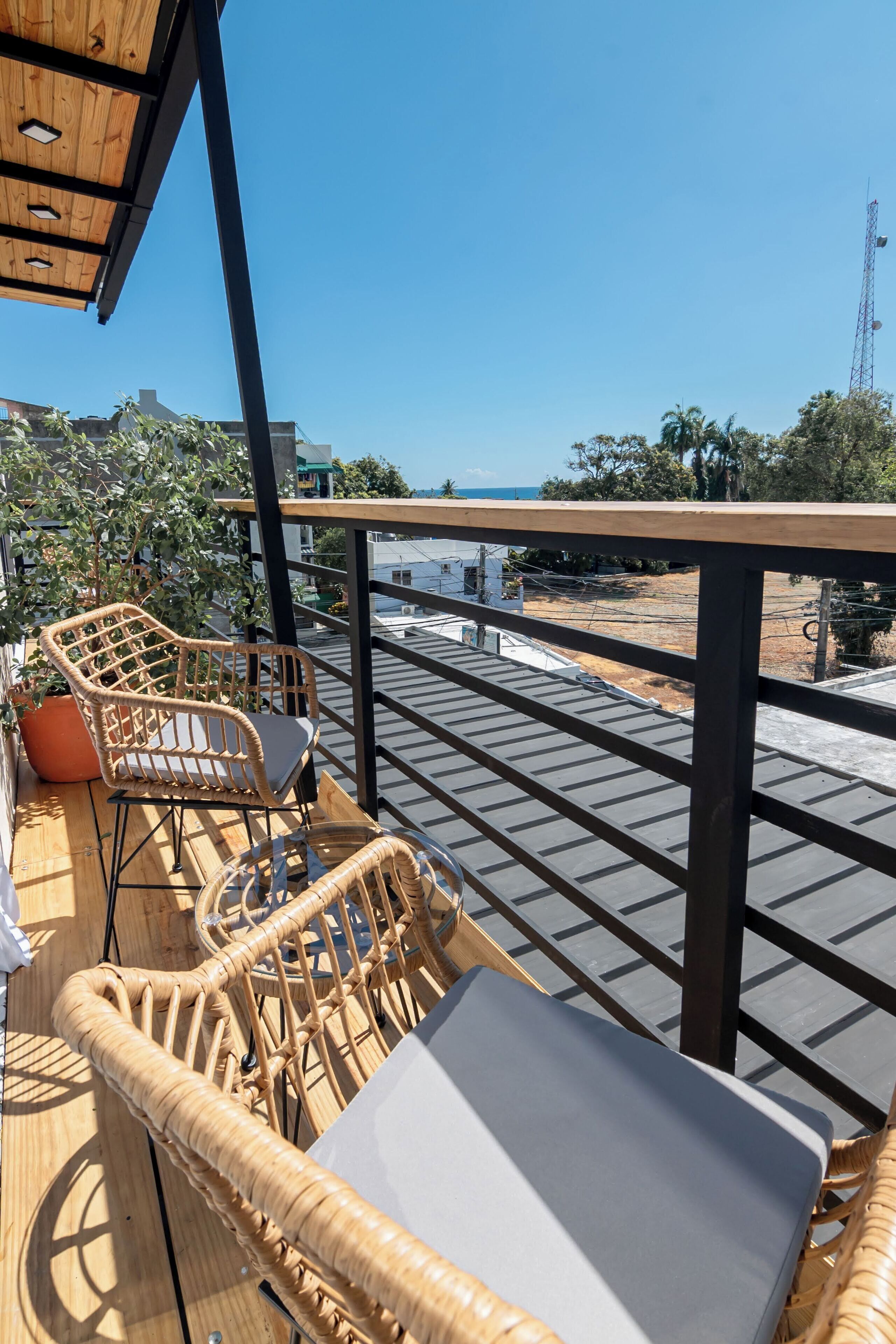 A balcony with wicker furniture, a potted plant, and a view of the sea and buildings.