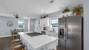 A modern kitchen with white cabinetry, a stainless steel refrigerator, and a central island with stools. - Modern Duplex, Sleeps 10, Dog Friendly + Fenced Yard, Full Kitchen & Kitchenette (Galveston)