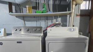 Duas máquinas de lavar Maytag sobre uma plataforma de madeira, com uma prateleira de metal acima contendo uma garrafa verde e uma amarela.