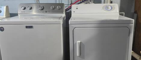 Two Maytag washing machines on a wooden platform with a metal shelf above them, containing a green bottle and a yellow bottle.