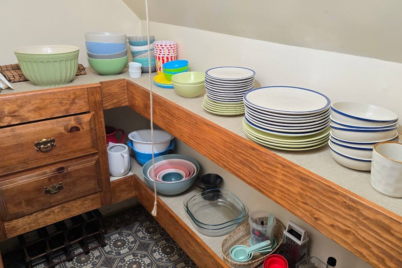 A kitchen with wooden cabinets, a countertop with stacked plates, bowls, and a hanging rack.