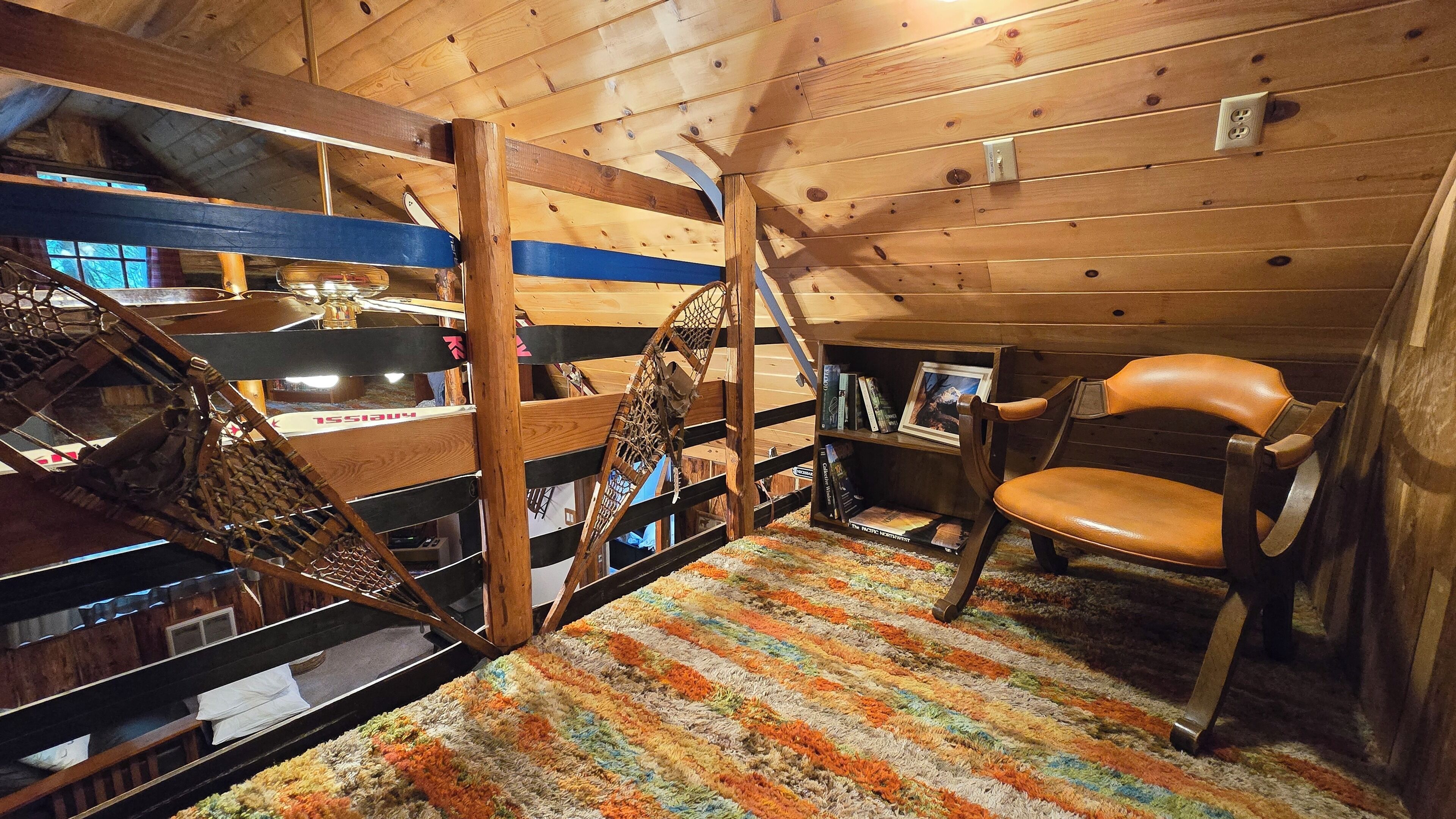 A cozy attic room with a wooden ceiling, a hammock, a chair, and a bookshelf.